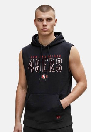 Młody mężczyzna z krótkimi włosami w czarnej bezrękawnikowej bluzie z kapturem z logo i napisem San Francisco 49ers, z jedną ręką w przedniej kieszeni.