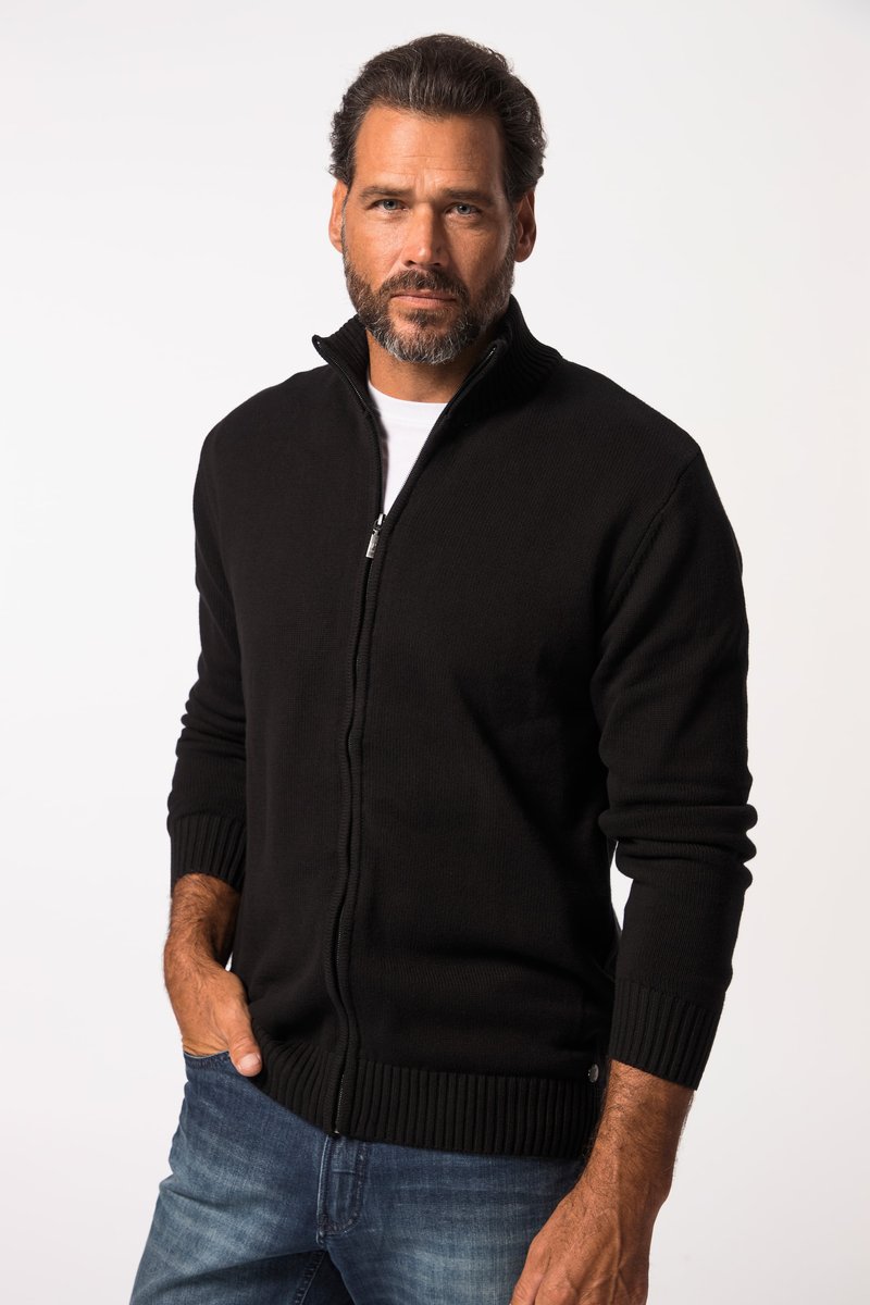 Homme avec une barbe portant un pull noir zippé sur une chemise blanche et un jean bleu, debout avec une main dans la poche.