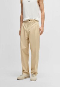 Beige wide-leg trousers with a high waist, front pleats, and a single button closure, paired with light sneakers and a white tank top.