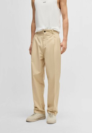 Beige weit geschnittene Hosen mit hoher Taille, vorderen Falten und einem einzigen Knopfverschluss, kombiniert mit hellen Sneakers und einem weißen Tanktop.