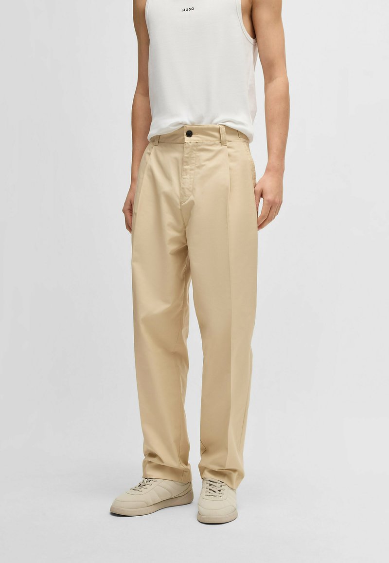 Beige wide-leg trousers with a high waist, front pleats, and a single button closure, paired with light sneakers and a white tank top.