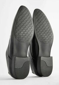 Zapatos de cuero negros exhibidos desde la parte inferior, con suelas de goma texturada con un patrón en forma de abanico y secciones de talón reforzadas.