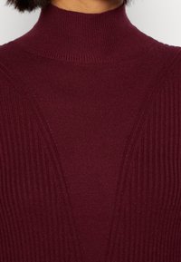 Pull col roulé bordeaux présentant une texture côtelée et des lignes de design diagonales, fabriqué en tricot doux et extensible.