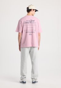 Light pink cotton t-shirt with short sleeves, featuring “New York Tennis Society” graphic and court outline on the back, paired with gray sweatpants.