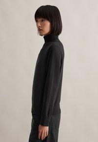 Fiatal nő egyenes fekete hajjal, fekete garbó pulóverben és sötét nadrágban, profilban áll, semleges háttér előtt.