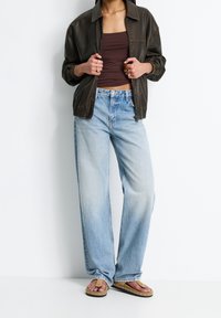 Personne portant une veste en cuir marron foncé, un haut marron, un jean évasé bleu clair et des sandales à boucle en cuir beige avec des clous.