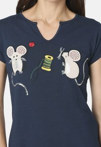 Una camiseta de color azul marino presenta ratones bordados y un hilo de costura amarillo. El diseño incluye acentos rojos y metálicos juguetones en la tela.