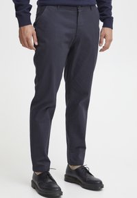 Marineblaues, schmal geschnittenes Tuchhose, gefertigt aus einem glatten Stoff. Mit Fronttaschen und Knopfverschluss, kombiniert mit schwarzen Schnürschuhen.