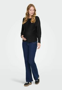 Maglione nero lavorato a maglia con scollo ampio e maniche a campana, abbinato a jeans svasati blu scuro. La modella ha una leggera curvatura alla vita.