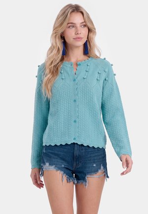 Lyshåret kvinde iført blå kvastøreringe, lyseblå tekstureret cardigan med knapper og pomponer og slidte denimshorts, stående mod en hvid baggrund.