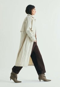 Kvinde iført beige trenchcoat, mørkebrune manchetbukser og spidse hælede støvler, gående på en ensartet lys baggrund.