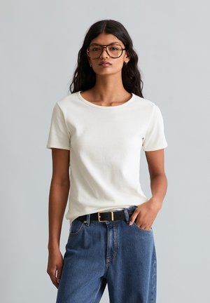 Frau mit langen dunklen Haaren, die große Brillen, ein weißes kurzärmeliges Oberteil, blaue Jeans und einen schwarzen Gürtel trägt und mit einer Hand in der Tasche steht.