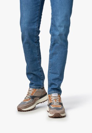 Mann, der schmale blaue Jeans trägt und braune, beige sowie schwarze Sneakers mit strukturierten Sohlen, auf einer einfachen weißen Fläche steht.