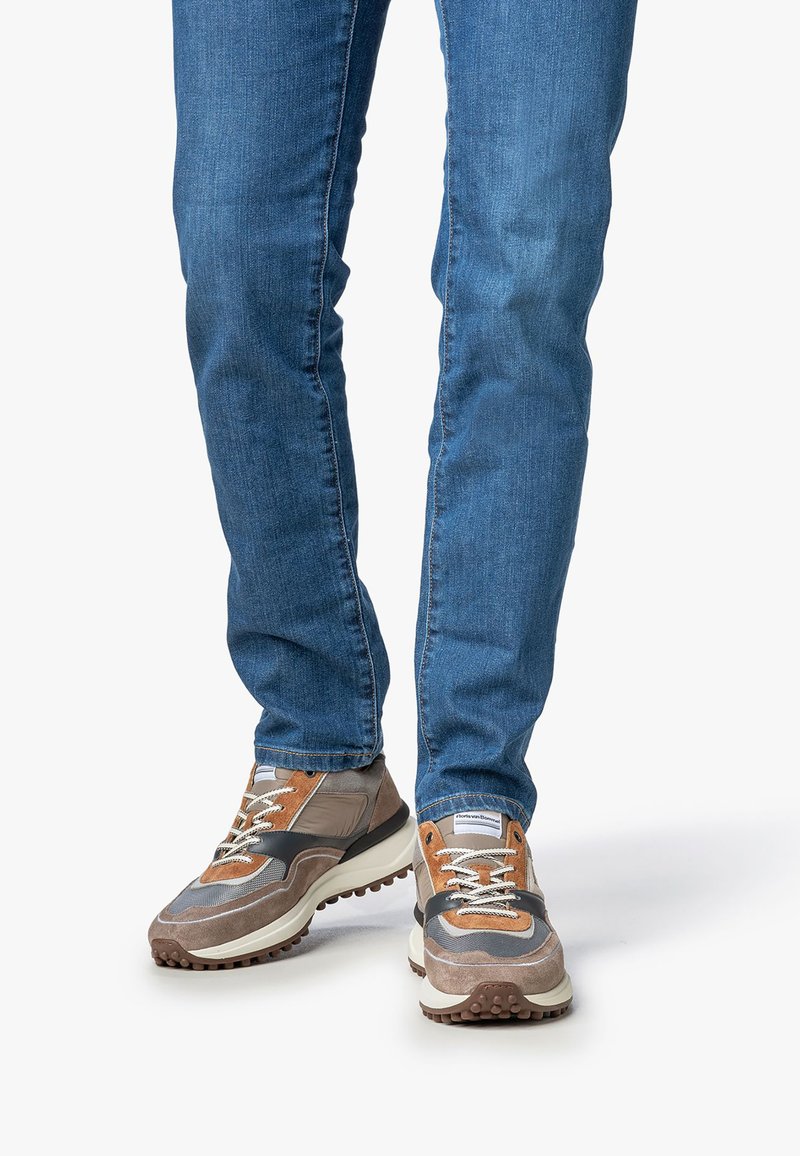 Uomo che indossa jeans slim blu e sneakers marroni, beige e nere con suole testurizzate, in piedi su una superficie bianca uniforme.