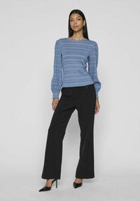 Modrá dlouhá halenka s texturovanými pruhy, doplněná o černé kalhoty s vysokým pasem a širokými nohavicemi a černé lodičky s špičatou špičkou.