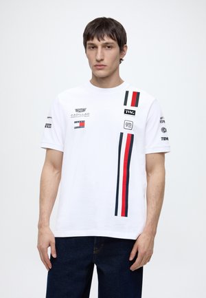 Jeune homme portant un t-shirt blanc au thème de course avec des rayures verticales rouges, blanches et bleues et plusieurs logos de marques, debout devant un fond uni.