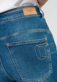 Jeans de denim azul con una etiqueta de cuero marrón en la cinturilla, costura decorativa en el bolsillo trasero y un corte clásico con textura suave.