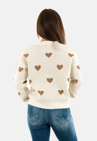 Donna con lunghi capelli castani, che indossa un maglione color crema con motivi di cuori marroni e jeans blu, in piedi con le mani sui fianchi e rivolta di spalle.