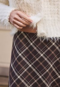 Pull blanc en tricot duveteux avec bordure à franges, associé à une jupe à carreaux foncée comportant des lignes marron, blanches et violet clair.