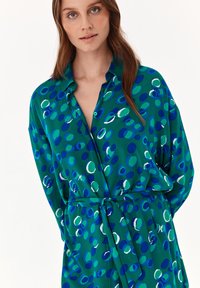 Abito verde a camicia con vita legata, caratterizzato da un mix di motivi circolari blu e bianchi. Realizzato in un tessuto morbido con una vestibilità rilassata.