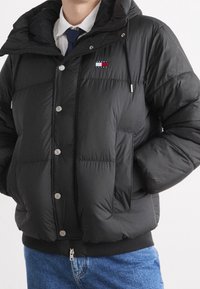 Schwarze gepolsterte Jacke mit Reißverschluss und Druckknopfverschluss, ausgestattet mit einer Kapuze, Fronttaschen und einem kleinen Logo-Patch auf der Brust. Weicher Stoff.