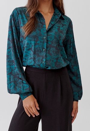 Woman wearing a teal and black floral long-sleeve button-up blouse tucked into high-waisted black pants.