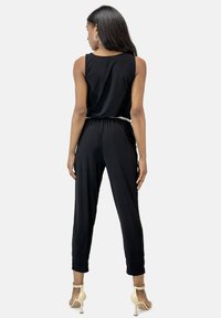 Mono negro con diseño sin mangas, cintura elástica y un cinturón decorativo. Tejido suave y piernas cónicas. Llevado con tacones marrón claro.