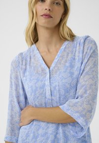 Light blue blouse with a floral pattern, sheer texture, three-quarter sleeves, v-neckline, and button detail. Soft, lightweight fabric.