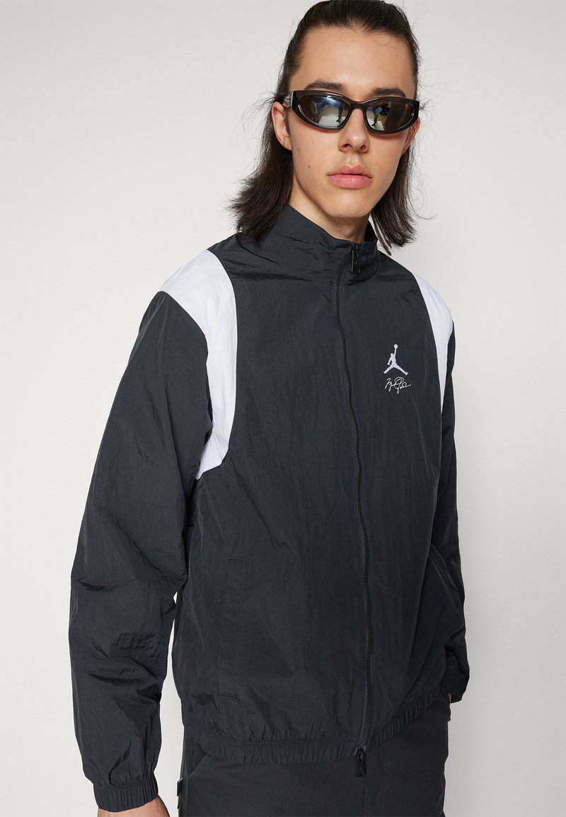 Veste coupe-vent noir et blanc avec fermeture éclair sur le devant, présentant un logo sur la poitrine et des poignets élastiques. Texture légère et froissée.
