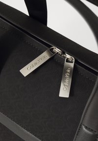 Sort lynlås taske med en struktureret overflade, der har et geometrisk mønster. Sølvfarvede detaljer inkluderer to mærker med "Calvin Klein" indgraveret.