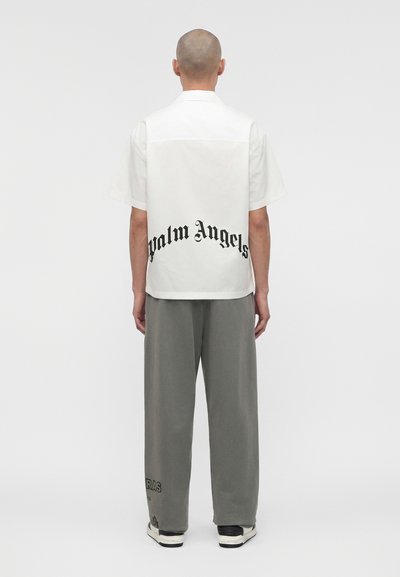 Άτομο με φαλάκρα στέκεται με την πλάτη προς την κάμερα, φοράει λευκό κοντομάνικο πουκάμισο με το κείμενο «Palm Angels» στην πλάτη και χαλαρό γκρι παντελόνι με μαύρα αθλητικά παπούτσια.