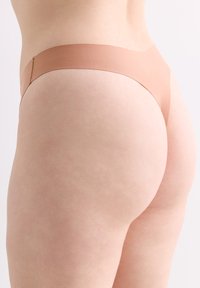 Nude nahtloser Tanga aus weichem Material, ausgestattet mit einem breiten elastischen Bund und minimaler Abdeckung, entworfen für Komfort und eine glatte Passform.