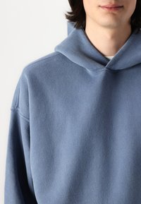 Sweat-shirt à capuche bleu avec une texture douce, offrant une coupe décontractée et un large col. Le tissu semble être en polaire.