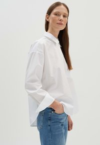 Camicetta bianca a maniche lunghe con vestibilità morbida, dotata di colletto e ampi polsini, abbinata a jeans blu. Texture liscia e design minimalista.