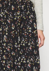 Jupe plissée noire avec un petit imprimé floral blanc, jaune et rouge, associée à une manche de pull en maille côtelée beige et une main visible sur le côté.