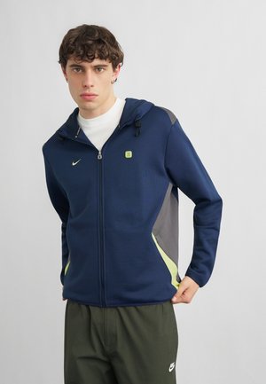 Joven con sudadera con capucha azul marino con cremallera y detalles neón y pantalones Nike verde oliva, mirando directamente hacia adelante con expresión neutral.