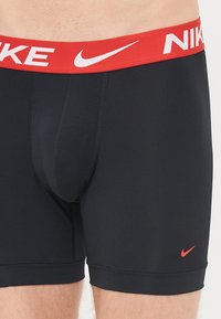 Boxeri atletici negri cu un elastic roșu la talie pe care se află logo-ul Nike. Materialul pare neted și aderent. Logo mic roșu aproape de tiv.