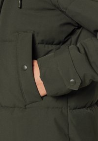Mørkegrønn dunjakke med sidelomme og trykknapp-lukking. Har en glatt stoffstruktur og skreddersydde mansjett detaljer.