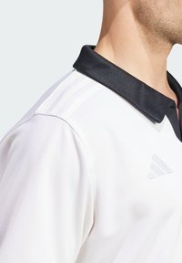 Hvid polo shirt med en sort krave, prydet med tre hvide striber på skulderen og et sølvlogo på brystet. Glat stofstruktur.