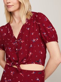 Burgundy blus med kort puffärm, blommönster i rött, blått och vitt, V-ringning och knuten detalj fram. Enfärgad baksida.