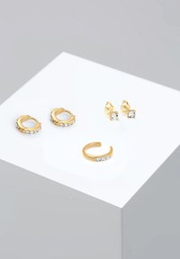 Conjunto de pendientes en tono dorado, que incluye dos pares de aros con piedras transparentes y un solo pendiente tipo cuff con tres piedras transparentes.