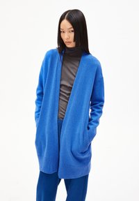 Blauwe open voorkardigan met lange mouwen, gebreide textuur, twee voorzakken, gedragen over een donkere coltrui en bijpassende broek.