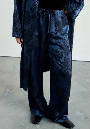 Pantalon en soie bleu foncé et longue veste assortie avec des motifs floraux noirs, portés avec des chaussures noires à bout carré.