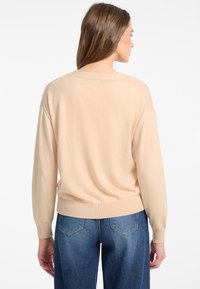 Šviesiai persiko spalvos megztinis su laisvu siluetu, plačiu iškirpimu ir raukšlėtu apvadu, derinamas su mėlynomis aukštu liemeniu džinsais.