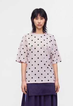 Jeune femme aux cheveux noirs portant un T-shirt rose clair oversize à pois noirs et une jupe bleu foncé, debout sur un fond blanc.