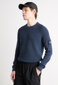 Maglione a maglia navy con texture a coste, scollo rotondo e patch del marchio ricamato sulla manica. Indossato con pantaloni neri con coulisse.