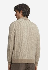 Beige stickad tröja med ribbad textur, som har en krage och manschettkantade nederkanter. Enkel design som framhäver värme och komfort.