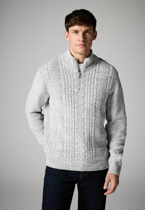 Sivi pleteni pulover s visokim ovratnikom, s teksturiranim kablovskim uzorcima i zatvaranjem na polu-zip. Nosi se preko tamnih traperica.