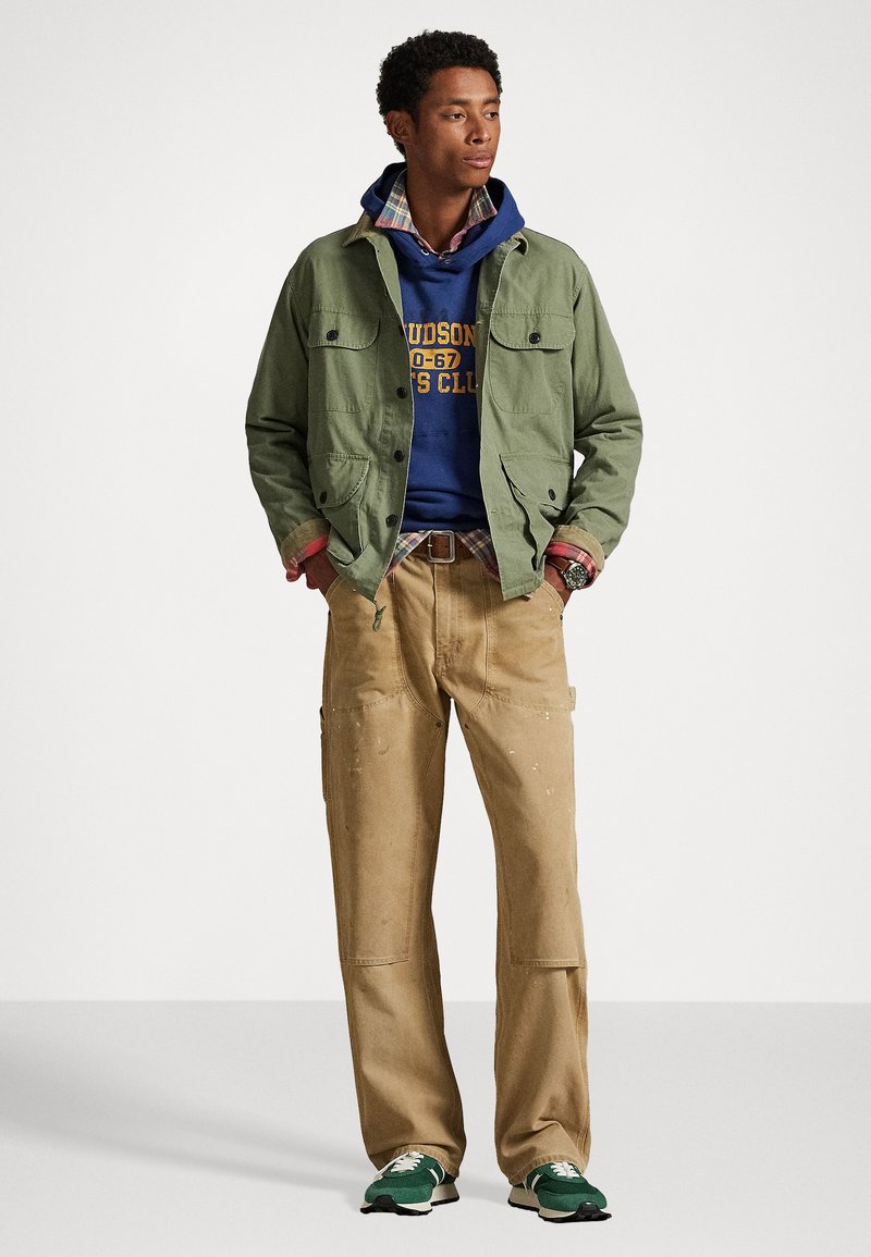 Giacca in tela verde, felpa con cappuccio blu, camicia a quadri sotto, pantaloni cargo beige e sneakers verdi. Design semplice, stile utilitario.
