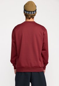 Sweatshirt bordeaux à manches longues, encolure ronde et poignets côtelés. Texture lisse et coupe décontractée. La vue arrière montre un design simple.